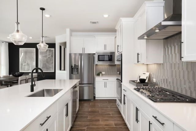 kitchen featuring wall chimney range hood, sink, appliances with stainless steel finishes, decorative light fixtures, and white cabinetry