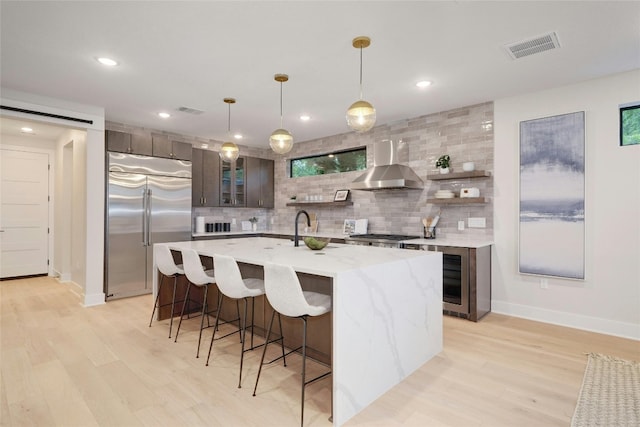 kitchen featuring pendant lighting, built in refrigerator, wall chimney exhaust hood, an island with sink, and dark brown cabinets