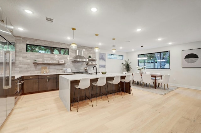 kitchen featuring decorative light fixtures, light hardwood / wood-style flooring, wall chimney exhaust hood, tasteful backsplash, and dark brown cabinets