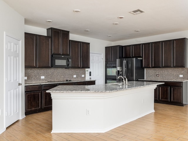 kitchen featuring an island with sink, light wood-type flooring, black appliances, and dark brown cabinetry