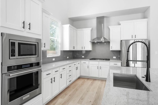 kitchen featuring backsplash, light stone counters, wall chimney exhaust hood, stainless steel appliances, and white cabinets