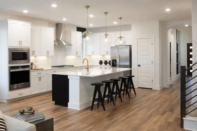 kitchen featuring white cabinets, hanging light fixtures, wall chimney exhaust hood, an island with sink, and stainless steel appliances