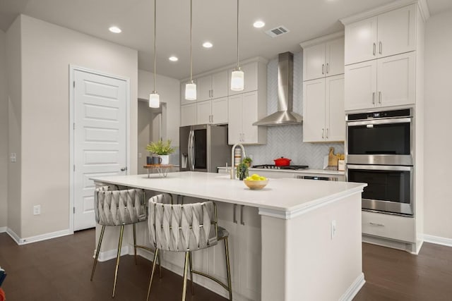 kitchen with stainless steel appliances, wall chimney range hood, an island with sink, decorative light fixtures, and white cabinets