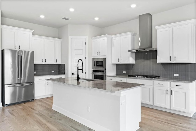 kitchen with sink, wall chimney range hood, a center island with sink, white cabinets, and appliances with stainless steel finishes