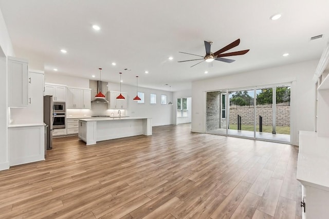 kitchen with white cabinets, hanging light fixtures, wall chimney exhaust hood, ceiling fan, and an island with sink