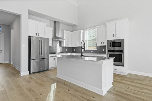 kitchen with white cabinets, wall chimney range hood, stainless steel appliances, and an island with sink