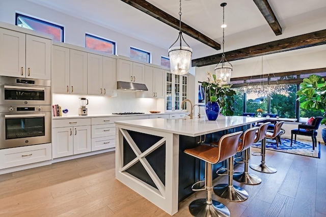 kitchen featuring double oven, decorative light fixtures, beamed ceiling, white cabinets, and an island with sink