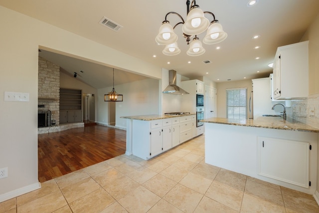 kitchen with white cabinets, wall chimney range hood, sink, a fireplace, and decorative light fixtures