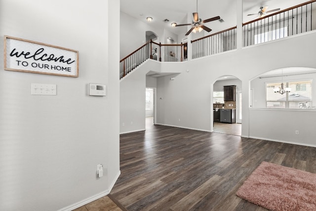 unfurnished living room with a towering ceiling, a wealth of natural light, ceiling fan with notable chandelier, and dark wood-type flooring