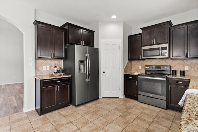 kitchen with dark brown cabinetry, stainless steel appliances, decorative backsplash, light stone counters, and light tile patterned floors