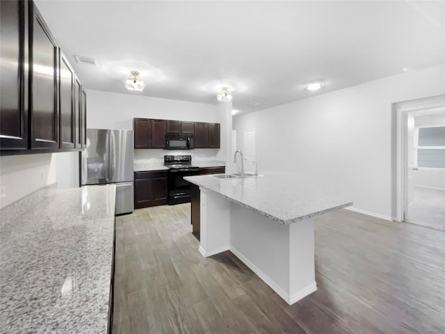 kitchen with light stone countertops, dark brown cabinetry, sink, black appliances, and a center island with sink