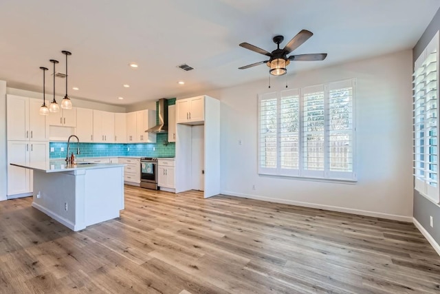 kitchen with tasteful backsplash, stainless steel electric stove, wall chimney range hood, white cabinetry, and an island with sink