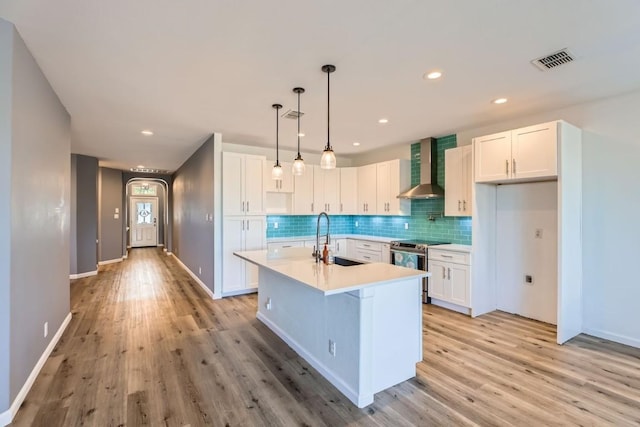 kitchen featuring wall chimney exhaust hood, stainless steel electric stove, sink, white cabinetry, and an island with sink