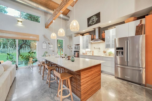 kitchen featuring wall chimney range hood, hanging light fixtures, a towering ceiling, white cabinetry, and stainless steel appliances