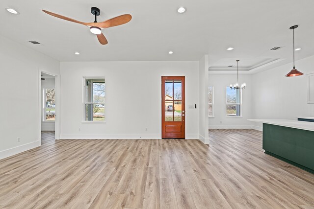 unfurnished living room featuring ceiling fan with notable chandelier, a tray ceiling, and light hardwood / wood-style flooring