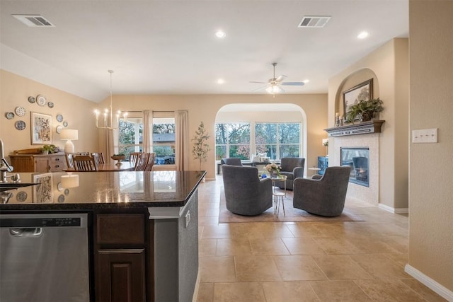 kitchen featuring dishwasher, a tiled fireplace, ceiling fan with notable chandelier, dark stone counters, and pendant lighting