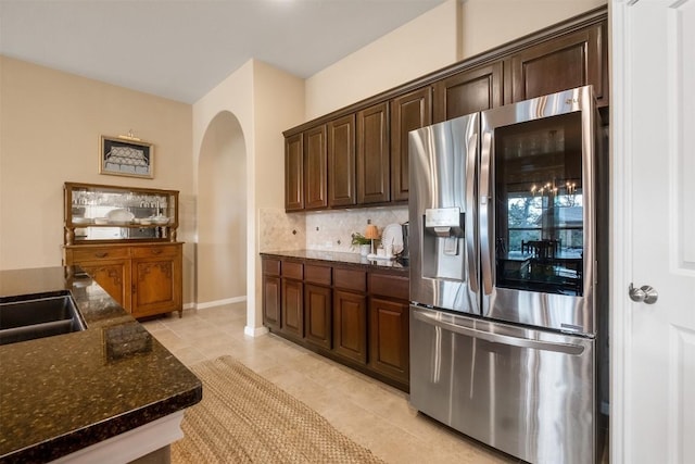 kitchen featuring light tile patterned flooring, backsplash, dark stone counters, dark brown cabinetry, and stainless steel fridge