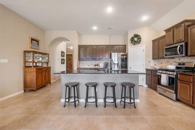 kitchen with sink, dark stone countertops, an island with sink, backsplash, and appliances with stainless steel finishes