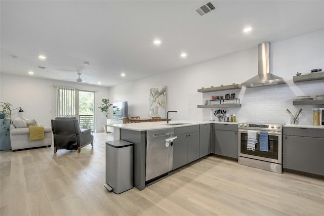 kitchen with ceiling fan, sink, stainless steel appliances, wall chimney range hood, and light wood-type flooring