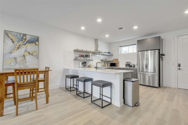 kitchen featuring a kitchen breakfast bar, wall chimney exhaust hood, light hardwood / wood-style floors, kitchen peninsula, and stainless steel refrigerator