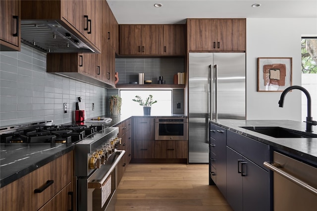 kitchen with dark stone counters, premium appliances, sink, exhaust hood, and light hardwood / wood-style flooring