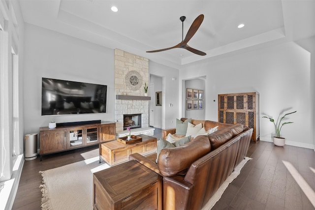 living room with a tray ceiling, a stone fireplace, dark hardwood / wood-style floors, and ceiling fan