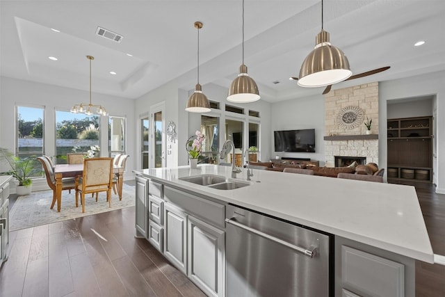 kitchen with stainless steel dishwasher, a raised ceiling, sink, a center island with sink, and a fireplace