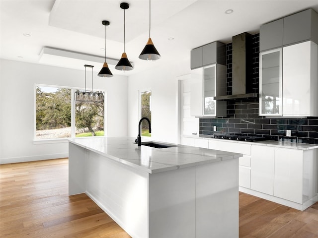 kitchen featuring light stone countertops, wall chimney range hood, sink, hanging light fixtures, and an island with sink