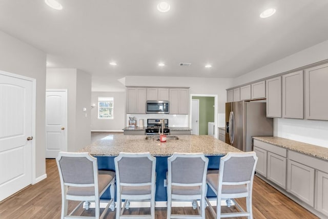 kitchen with light stone countertops, appliances with stainless steel finishes, light wood-type flooring, a kitchen island with sink, and gray cabinets