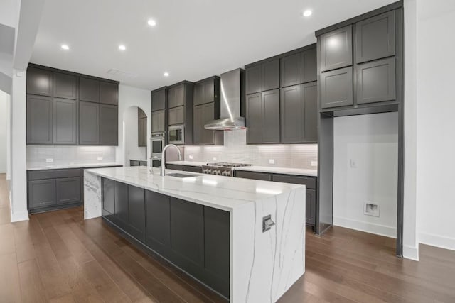 kitchen featuring sink, wall chimney exhaust hood, light stone countertops, an island with sink, and dark hardwood / wood-style flooring