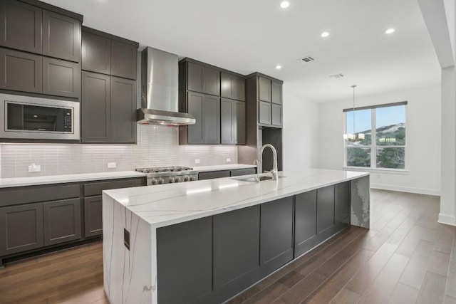 kitchen with sink, wall chimney exhaust hood, light stone counters, an island with sink, and appliances with stainless steel finishes