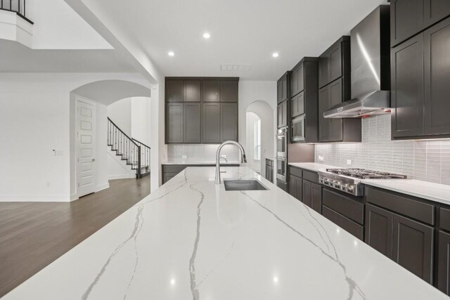 kitchen with wall chimney range hood, sink, decorative backsplash, light stone counters, and stainless steel appliances