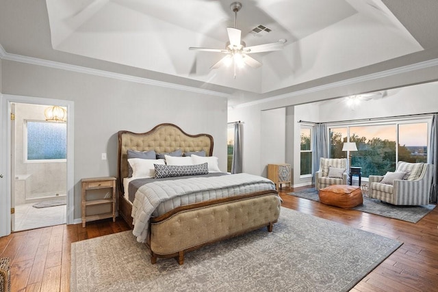 bedroom featuring wood finished floors, visible vents, a raised ceiling, ensuite bath, and crown molding