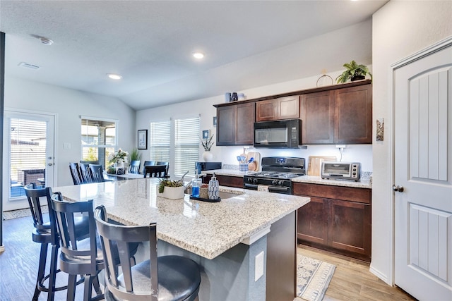 kitchen featuring black appliances, a kitchen bar, an island with sink, dark brown cabinets, and light stone counters