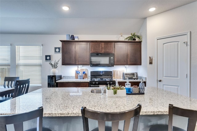 kitchen with a center island with sink, black appliances, a kitchen breakfast bar, dark brown cabinets, and light stone counters