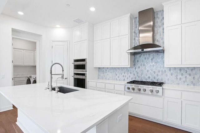 kitchen with a center island with sink, stainless steel appliances, a sink, wall chimney range hood, and independent washer and dryer