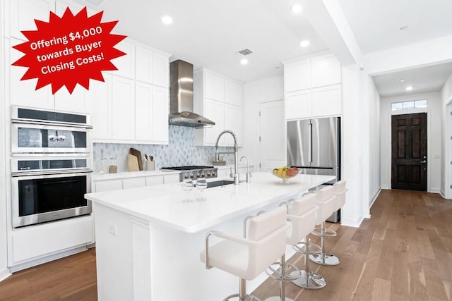 kitchen featuring white cabinets, stainless steel appliances, a center island with sink, light wood-type flooring, and wall chimney exhaust hood