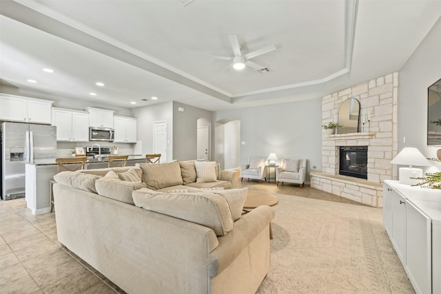 living room featuring a tray ceiling, a stone fireplace, ceiling fan, and light tile patterned floors