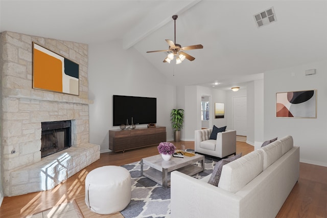 living room with ceiling fan, wood-type flooring, a stone fireplace, and lofted ceiling with beams