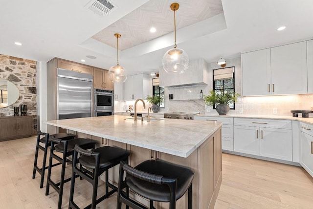 kitchen with white cabinets, hanging light fixtures, a kitchen island with sink, a raised ceiling, and light stone countertops