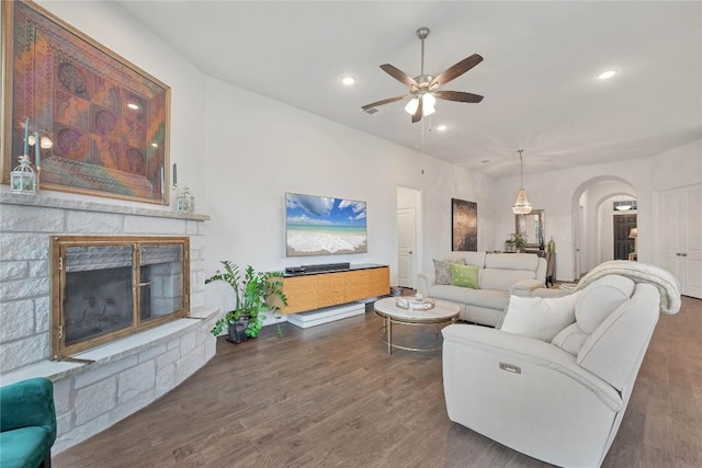 living room featuring dark wood-type flooring, ceiling fan, and a stone fireplace