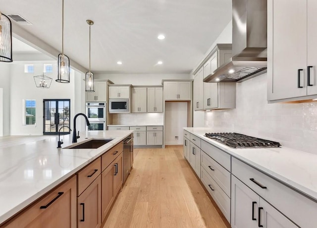 kitchen with light stone countertops, wall chimney range hood, pendant lighting, appliances with stainless steel finishes, and light wood-type flooring