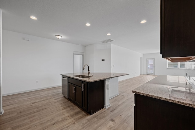 kitchen with a kitchen island with sink, sink, stainless steel dishwasher, light wood-type flooring, and light stone countertops