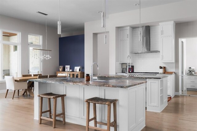 kitchen featuring pendant lighting, white cabinets, wall chimney range hood, decorative backsplash, and an island with sink