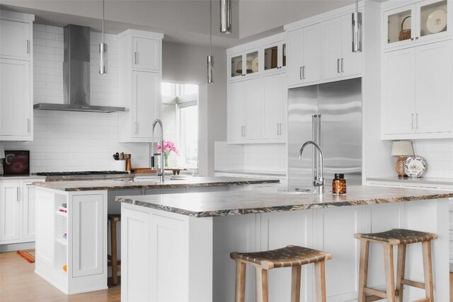 kitchen with stainless steel appliances, wall chimney range hood, decorative light fixtures, a breakfast bar area, and a center island with sink