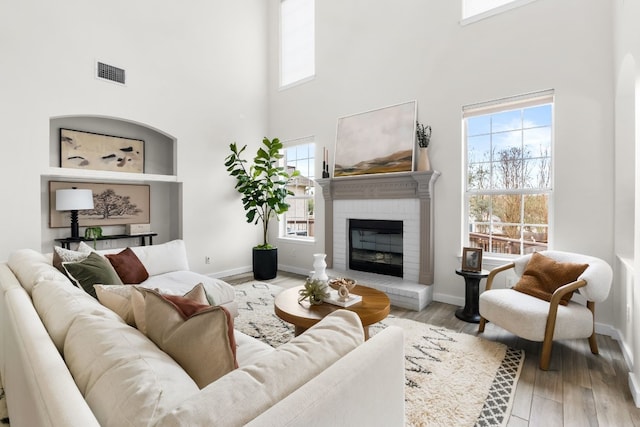 living room with light hardwood / wood-style floors, a towering ceiling, a healthy amount of sunlight, and a brick fireplace