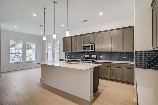 kitchen with sink, hanging light fixtures, stainless steel appliances, a kitchen island with sink, and light wood-type flooring