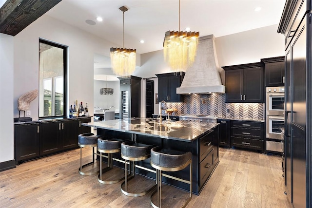 19204 Secretariat Pl, Spicewood, TX 78669 - kitchen with custom exhaust hood, light wood-style flooring, tasteful backsplash, stainless steel double oven, and an island with sink