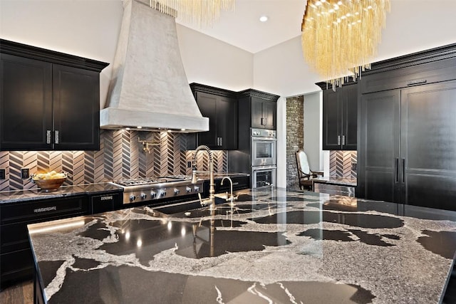 19204 Secretariat Pl, Spicewood, TX 78669 - kitchen with custom range hood, dark cabinetry, a towering ceiling, appliances with stainless steel finishes, and backsplash