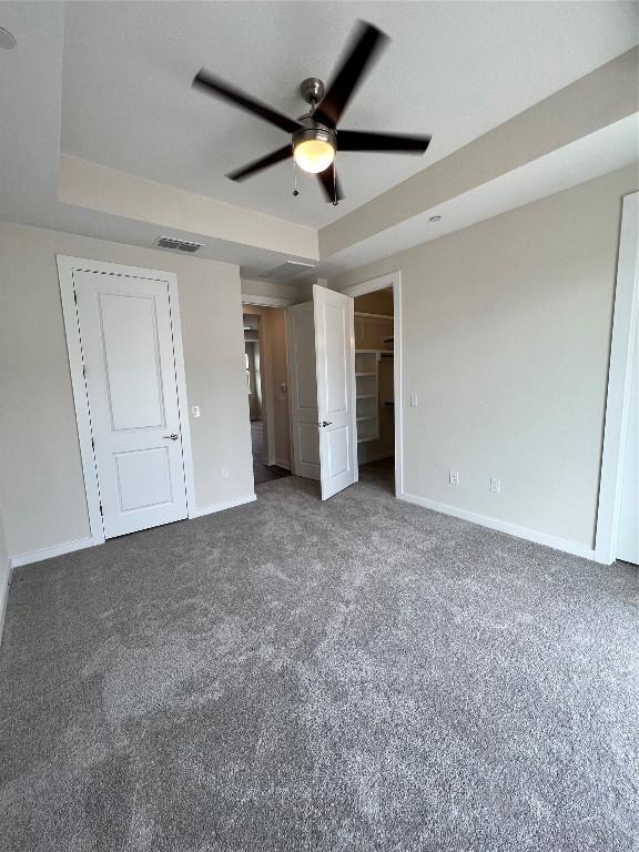 unfurnished bedroom with dark carpet, a tray ceiling, ceiling fan, and a spacious closet